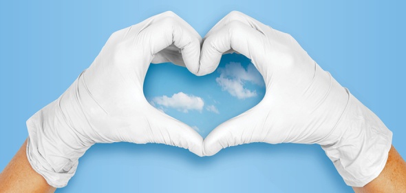 Photo of medical gloved hands making a heart shape against a blue sky.