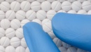 Background of round white pills with an indented line through the center. From the bottom right of the image a thumb and index finger in blue latex gloves extend holding another white pill with an indented line through the middle. 