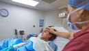 A brown haired woman lies in a hospital bed while in labor. Another woman in red scrbs, a medical mask, and a medical hairnet has her right hand on the other woman's head and her left hand below the covers. A hand in a green gown and a white medical glove reaches out from the bottom left frame and is placed on the woman in labor's abdomen. A clock on the far wall read 3:52. There is additional medical equipment around the room. 